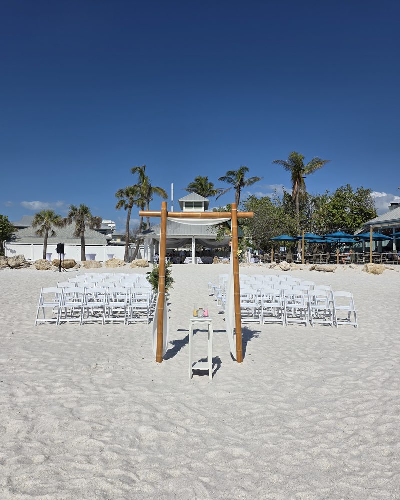 Beach wedding ceremony set up on white sand with a bamboo arch, draped fabric and rows of white chairs