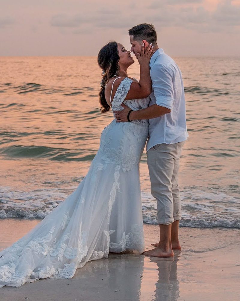Bride in a lace gown and groom kissing barefoot in the shallow surf at sunset on a Gulf Coast beach