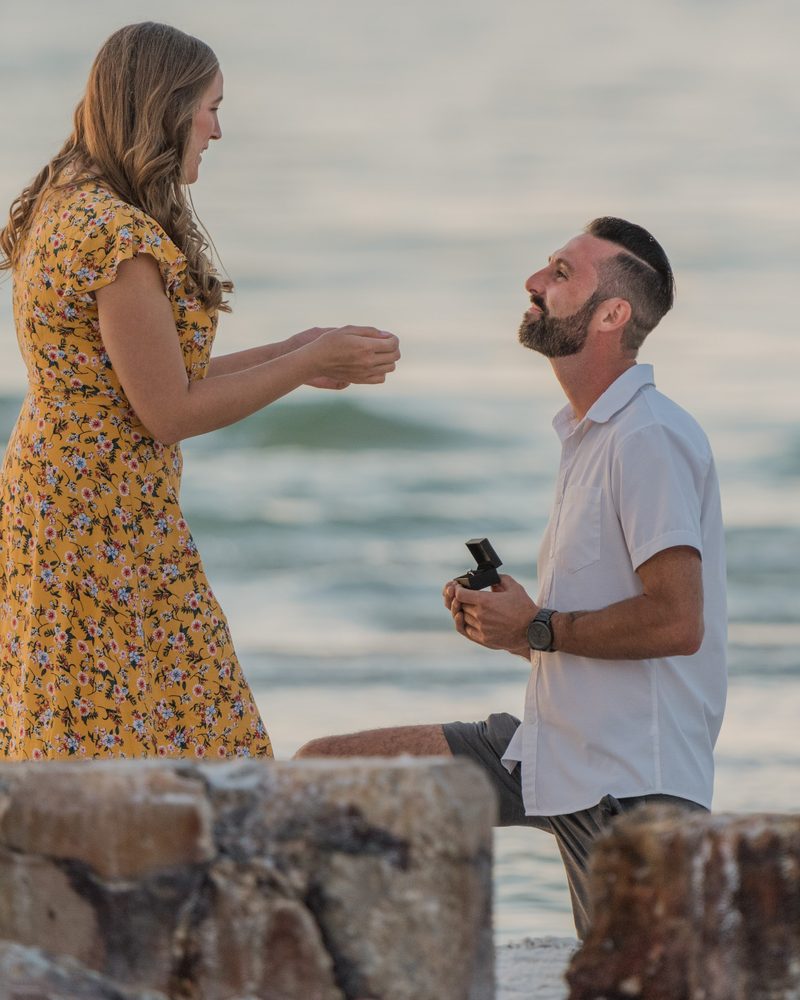 Man on one knee holding a ring box, proposing to his partner on the rocks at the water's edge