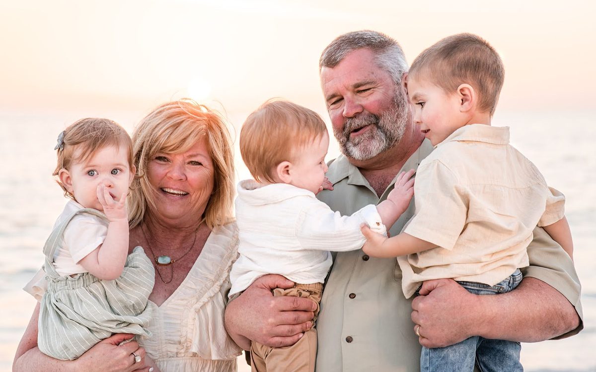 Grandparents laughing while holding three young grandchildren on the beach at sunset