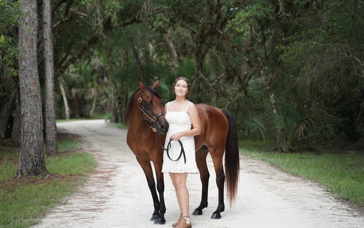 High school senior in a white dress standing with her horse on a sandy road under live oaks