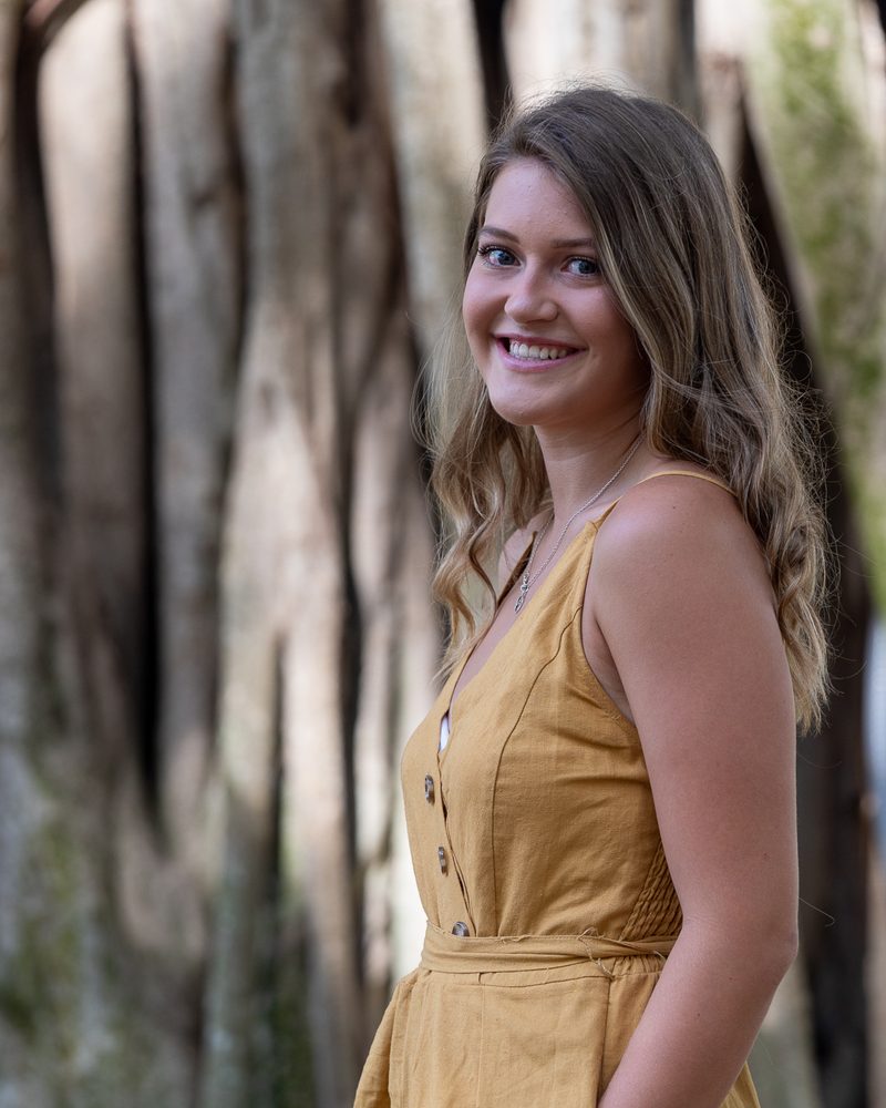High school senior in a mustard dress laughing in front of tall banyan tree roots